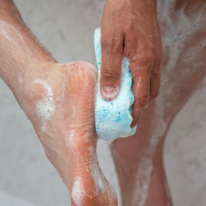 Person scrubbing foot with a blue sponge in a bathtub filled with water and soap suds.